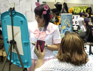 Brandi Miller, owner of the online business Dorky Dino and UCM alumna, works on a watercolor painting Saturday at her booth during the Kansas City Comic Con. Her friend, Andi Dieckman, an academic adviser in the College of Health, Science and Technology, holds up a cellphone with a picture of a horse for reference.